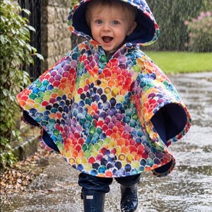 May include: A hooded poncho featuring a vibrant, multi-colored scale pattern in shades of red, orange, yellow, green, blue, and purple. The garment has a dark blue interior lining, a single front button closure, and is worn with dark blue rain boots.