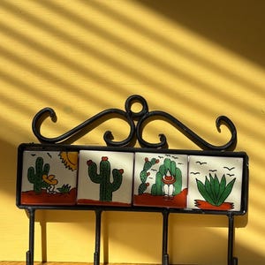 May include: A black metal wall hook rack featuring four square ceramic tiles with hand-painted desert scenes. Each tile displays green cacti, red ground, and a yellow sun or birds against a white background. The rack has a decorative scroll top.