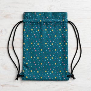 May include: A teal drawstring backpack featuring a repeating pattern of small yellow stars and pink and light blue dots. The bag has black cord straps that exit from the bottom corners and cinch the top opening closed.