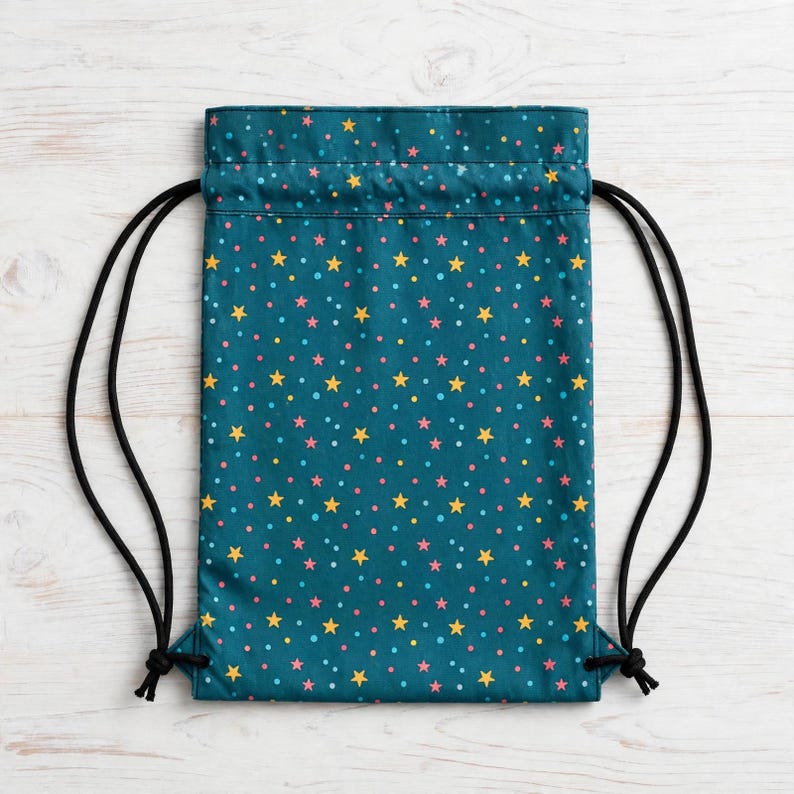 May include: A teal drawstring backpack featuring a repeating pattern of small yellow stars and pink and light blue dots. The bag has black cord straps that exit from the bottom corners and cinch the top opening closed.