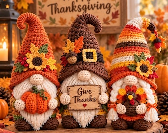 May include: Three crocheted gnomes with tall hats decorated for autumn. The left gnome holds an orange pumpkin, the centre gnome holds a sign reading Give Thanks, and the right gnome features braided hair and holds a small turkey figure.