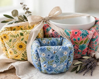 May include: Three quilted fabric bowl cozies featuring floral patterns in yellow, blue, and pink, tied together with a beige ribbon. The yellow cosy has sunflowers, the blue has small flowers and bees, and the pink has large floral prints.