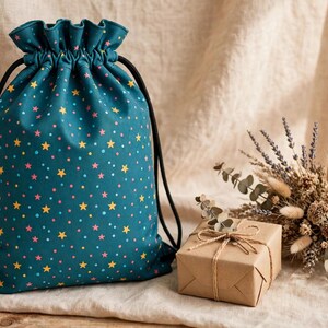 May include: A teal drawstring bag featuring a repeating pattern of small yellow stars and pink dots. The bag has a gathered top closure with black cord strings. Beside the bag sits a small square gift box wrapped in brown paper.