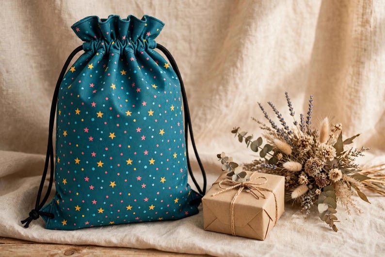 May include: A teal drawstring bag featuring a repeating pattern of small yellow stars and pink dots. The bag has a gathered top closure with black cord strings. Beside the bag sits a small square gift box wrapped in brown paper.