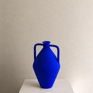 Puede incluir: Un jarrón de color azul cobalto vibrante con un cuerpo en forma de diamante y dos asas simétricas que se extienden desde el cuello hasta la sección media. El recipiente tiene un acabado mate y una abertura circular estrecha en la parte superior.