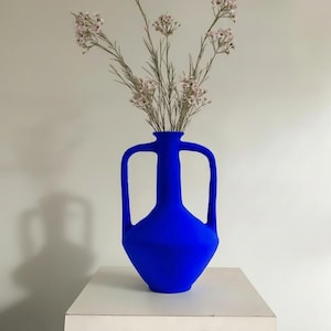 Puede incluir: Un jarrón de color azul cobalto vibrante con un cuerpo ancho y angular y dos asas simétricas que se extienden desde el cuello hasta el hombro. El jarrón contiene un ramo de delicadas flores secas de color claro con tallos finos y pequeñas flores.