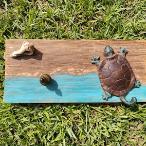 May include: A rustic wooden plaque with a brown and turquoise painted design, featuring a bronze turtle, a seashell, and a snail shell. The turtle has red accents on its head and is positioned on the right side of the plaque.