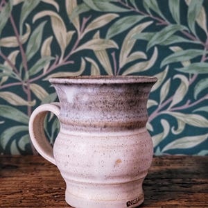 May include: A handcrafted ceramic mug with a speckled, pale pink body and a darker, grey-brown rim. The mug has a curved handle and a slightly flared top. The background features a green and cream leaf pattern.