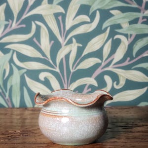 May include: A small, handmade ceramic bowl with a wavy edge. The bowl has a light grey body with a pale green band and a brown rim. The background is a teal wallpaper with a white and green leaf design.