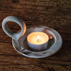 May include: A round, off-white ceramic candle holder with a speckled finish and a twisted loop handle. The holder features a shallow basin containing a small, lit tea light candle with a warm, glowing flame and a metallic silver casing.