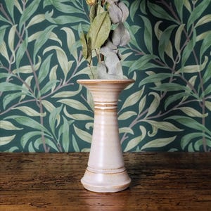 May include: A cream ceramic vase with a flared top and a wide base. The vase has a textured surface and brown accents. Dried flowers and leaves are arranged inside. The background features green and white floral wallpaper. The vase is likely 20 cm tall.