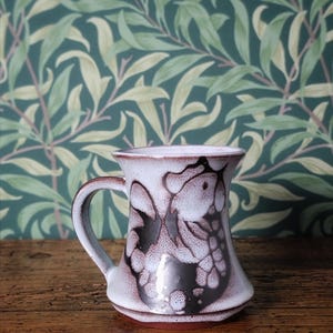 May include: A ceramic mug with a white and brown floral pattern. The mug features a handle and a flared base. The background is a green and white floral wallpaper. The mug rests on a wooden surface.