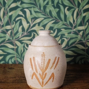 May include: A beige ceramic jar with a lid, decorated with an orange wheat stalk design. The jar is round and rests on a wooden surface. The background features green and white floral wallpaper.
