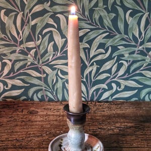 May include: A lit taper candle in a beige hue sits inside a ceramic candle holder. The holder features a wide, circular base with a speckled cream and brown glaze, decorated with a dark brown painted leaf pattern around the central stem.