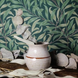 May include: A speckled ceramic vase with a brown stripe, holding dried eucalyptus branches. The vase is set on a wooden surface, with more eucalyptus leaves scattered around. The background features a green and cream leaf pattern.