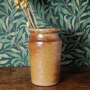 May include: A cylindrical, tan stoneware jar with a flared rim, containing dried yellow flowers. The rustic pot has a textured surface and sits on a wooden table. The background features a green and white leaf pattern.