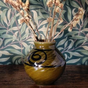 May include: An olive-green ceramic vase with a black spiral and stripe design, holding dried beige flowers. The vase is on a dark wooden surface, set against a patterned green and blue backdrop. The vase is approximately 15 cm tall.
