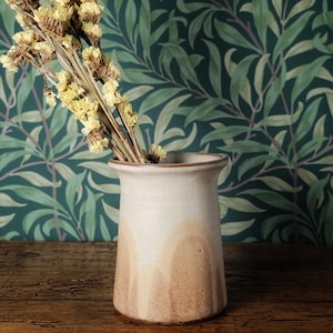 May include: A cream and brown ceramic vase with dried yellow flowers. The vase has a flared top and sits on a wooden surface. The background features a green and blue floral wallpaper. The vase is approximately 15 cm tall.