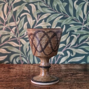 May include: A brown ceramic goblet featuring a speckled texture and a dark brown hand-painted lattice pattern around the bowl. The vessel has a flared rim, a slender stem with a central ring detail, and a circular base with a dark brown edge.