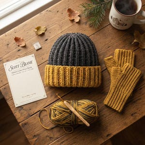 May include: A two-toned ribbed crochet beanie with a dark gray crown and a mustard yellow folded brim, accompanied by matching mustard yellow fingerless gloves, a ball of variegated yarn, a wooden crochet hook, and a product card labeled Scott Beanie.