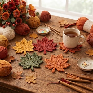 Puede incluir: Un conjunto de cinco posavasos de ganchillo con forma de hoja en colores amarillo, rojo, verde y naranja, dispuestos sobre una superficie de madera junto a ovillos de lana, ganchillos de madera, una taza de té y un ramo de flores de otoño.