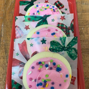 May include: Three frosted sugar cookies with pink icing and colorful sprinkles, presented in a red tray. The cookies sit on decorative paper with bows and stars. The cookies have a light yellow base.