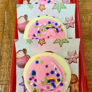 May include: A red tray holds frosted sugar cookies. Each cookie has a pink frosting swirl with colorful sprinkles. The cookies sit on paper with Christmas-themed illustrations, including snowflakes, gingerbread men, and hot cocoa mugs.