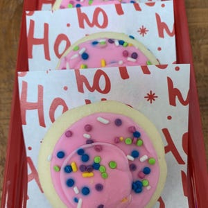 May include: A red tray holds three frosted sugar cookies. Each cookie has a pink frosting base with colorful sprinkles. The cookies rest on paper wrappers printed with "Ho" in red. The tray is on a wooden surface.