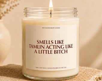 May include: A clear glass jar candle with a lit wick and cream-colored wax. The white label features the text THE SNOWDROP CANDLE, SMELLS LIKE TAMLIN ACTING LIKE A LITTLE BITCH, and 100% SOY WAX • HAND-POURED IN THE USA in dark brown lettering.
