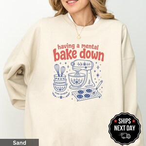 May include: A sand-colored crewneck sweatshirt featuring a graphic print in red and blue. The text reads "having a mental BAKE down" and "from scratch". The illustration includes a stand mixer, stacked bowls, a whisk, and cookies on a tray.