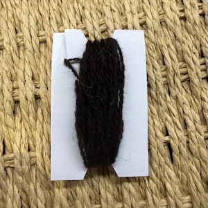 May include: A length of dark brown yarn wrapped around a white cardboard bobbin. The yarn has a textured, fibrous appearance and is wound vertically in multiple loops across the centre of the notched white paper card.
