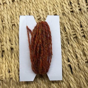 May include: A small bundle of variegated rust-orange and brown yarn wrapped around a white cardboard bobbin. The yarn features a textured, multi-tonal appearance with visible fibres, neatly wound in a vertical orientation on the card.