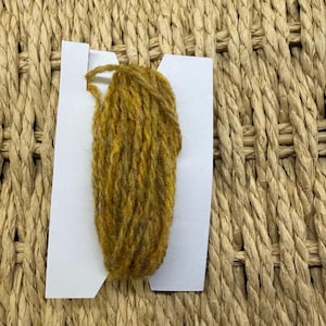 May include: A bundle of mustard yellow yarn wrapped around a white rectangular cardboard card. The yarn has a textured, fibrous appearance with visible strands wound vertically across the centre of the white card stock.