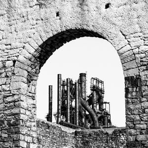 May include: A black and white photograph showing a weathered stone archway framing a complex industrial structure with tall metal pipes, cylindrical towers, and interconnected steel scaffolding, set against a bright, clear, and featureless sky.