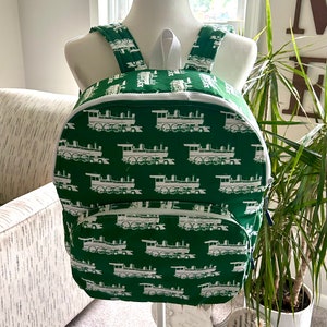 May include: A green backpack with a white train pattern. The backpack has a zippered pocket on the front and adjustable straps.