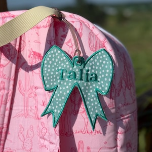 May include: A teal bow-shaped luggage tag with white polka dots and the name "Talia" embroidered in white. The tag is attached to a pink backpack with a cowboy and cactus pattern.