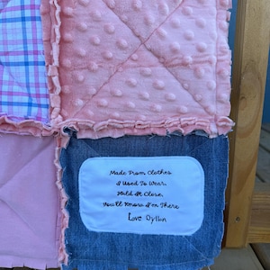 May include: A handmade patchwork quilt with various fabrics, including pink, blue, and denim. The quilt features a soft, textured pink fabric with a dotted pattern. A white patch with the text "Made From Clothes I Used To Wear...Love Dyllon" is sewn on the quilt.