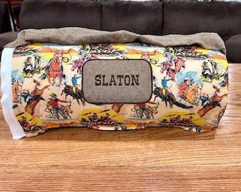 May include: A rectangular pillow with a cowboy-themed print featuring cowboys on horses, bucking broncos, and lassoing cattle. The pillow has a brown patch with the name "SLATON" embroidered on it. A tan fabric covers the top.