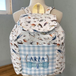 May include: A white backpack with a flap closure, featuring a pattern of colorful fishing flies. The front pocket has a blue and white gingham pattern with the name "ARZA" embroidered above two buckets and fish.
