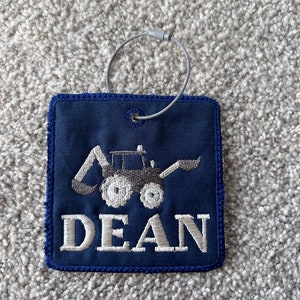 May include: A blue fabric key chain with a silver ring. The key chain has an embroidered image of a tractor and the name "DEAN" in white thread.