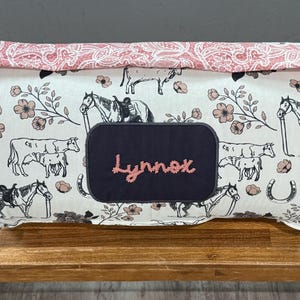 May include: A rectangular fabric storage bin with a white background featuring illustrations of horses, cows, and floral accents. The bin has a navy blue patch embroidered with the name "Lynnox" in pink.