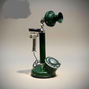 Może przedstawiać: Ciemnozielony telefon typu candlestick z chromowaną słuchawką, pasującą zieloną okrągłą tarczą numerową u podstawy oraz wyraźnym zielonym mikrofonem na górze. Urządzenie posiada czarny spiralny przewód łączący słuchawkę z podstawą.