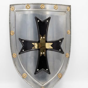 May include: A silver-colored metal heater shield featuring a prominent black cross with flared ends at the center. The shield is decorated with small gold-toned floral accents along the outer border and silver rivets throughout the design.