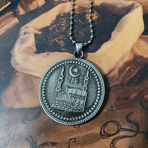 Pode incluir: Um colar com pingente circular de tom prateado apresentando um relevo de uma mesquita com dois minaretes, um símbolo de lua crescente e estrela, e o número 786 gravado no centro, suspenso por uma corrente de bolinhas prateada.