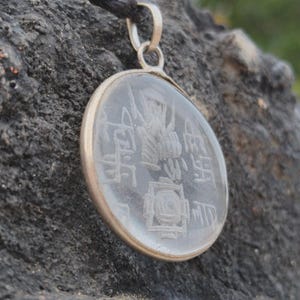 May include: A circular clear glass pendant set in a silver-toned metal frame, featuring etched white symbols and geometric patterns on its surface. The pendant is attached to a black cord, creating a necklace with a distinct, detailed design.