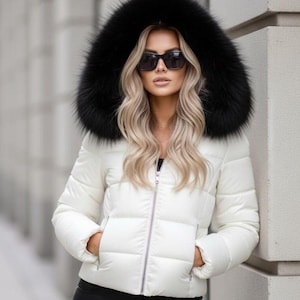 May include: A white puffer jacket featuring a large, black faux fur-trimmed hood, a central silver zipper closure, and two side pockets. The jacket is worn with black trousers and dark-tinted sunglasses, creating a high-contrast winter look.