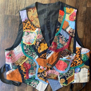 May include: A sleeveless vest constructed from a patchwork of various fabrics including floral prints, leopard patterns, and polka dots. The garment features a V-neckline, three front buttons, and decorative rickrack trim along the fabric seams.