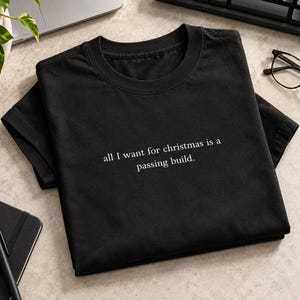 Peut inclure: Un t-shirt noir à manches courtes avec le texte imprimé en blanc "all I want for christmas is a passing build." Le t-shirt présente une encolure ronde et est posé à plat, mettant en valeur le texte centré sur le devant du vêtement.