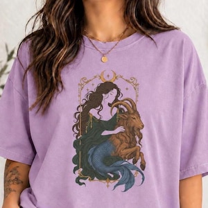 May include: A light purple t-shirt featuring a graphic illustration of a figure with long dark hair and a blue fish tail embracing a brown goat. The design is enclosed in a gold-colored ornate rectangular frame with a crescent moon at the top.