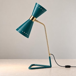 May include: A desk lamp featuring a dual-cone teal shade with small white circular perforations along the edges. The lamp has a polished gold-tone metal arm and a triangular teal base, connected by a black power cord with an inline switch.